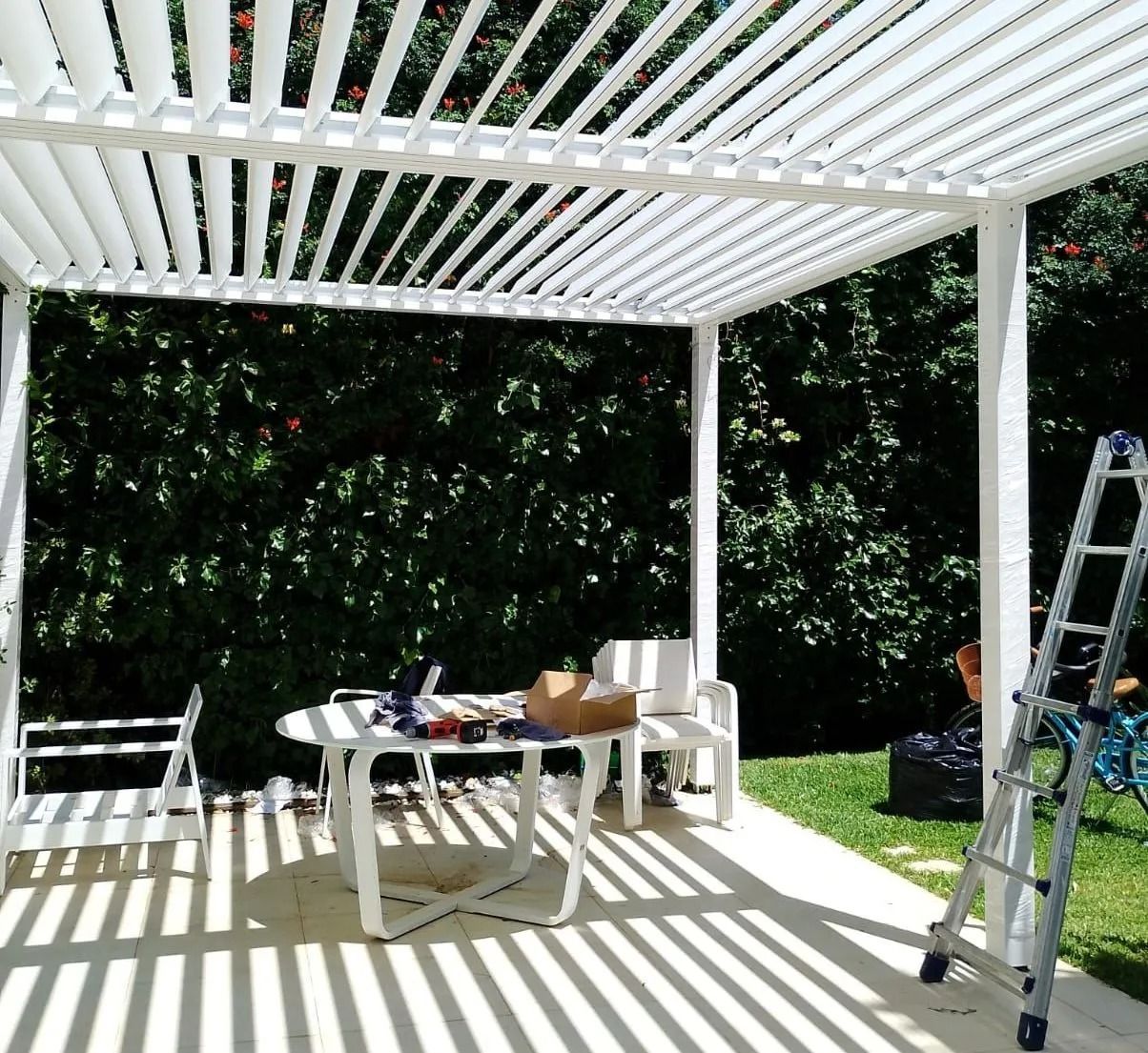Patio sul retro della casa sotto un pergolato bianco con tavolo, sedie, scaletta e luce del sole a terra