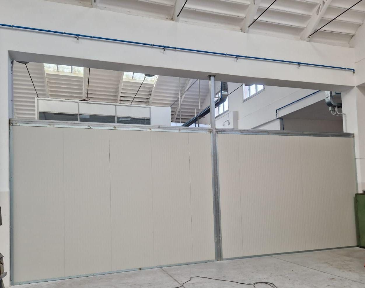 Cancello industriale beige chiuso all'interno di un magazzino con trave blu in alto e tetto in lamiera ondulata.