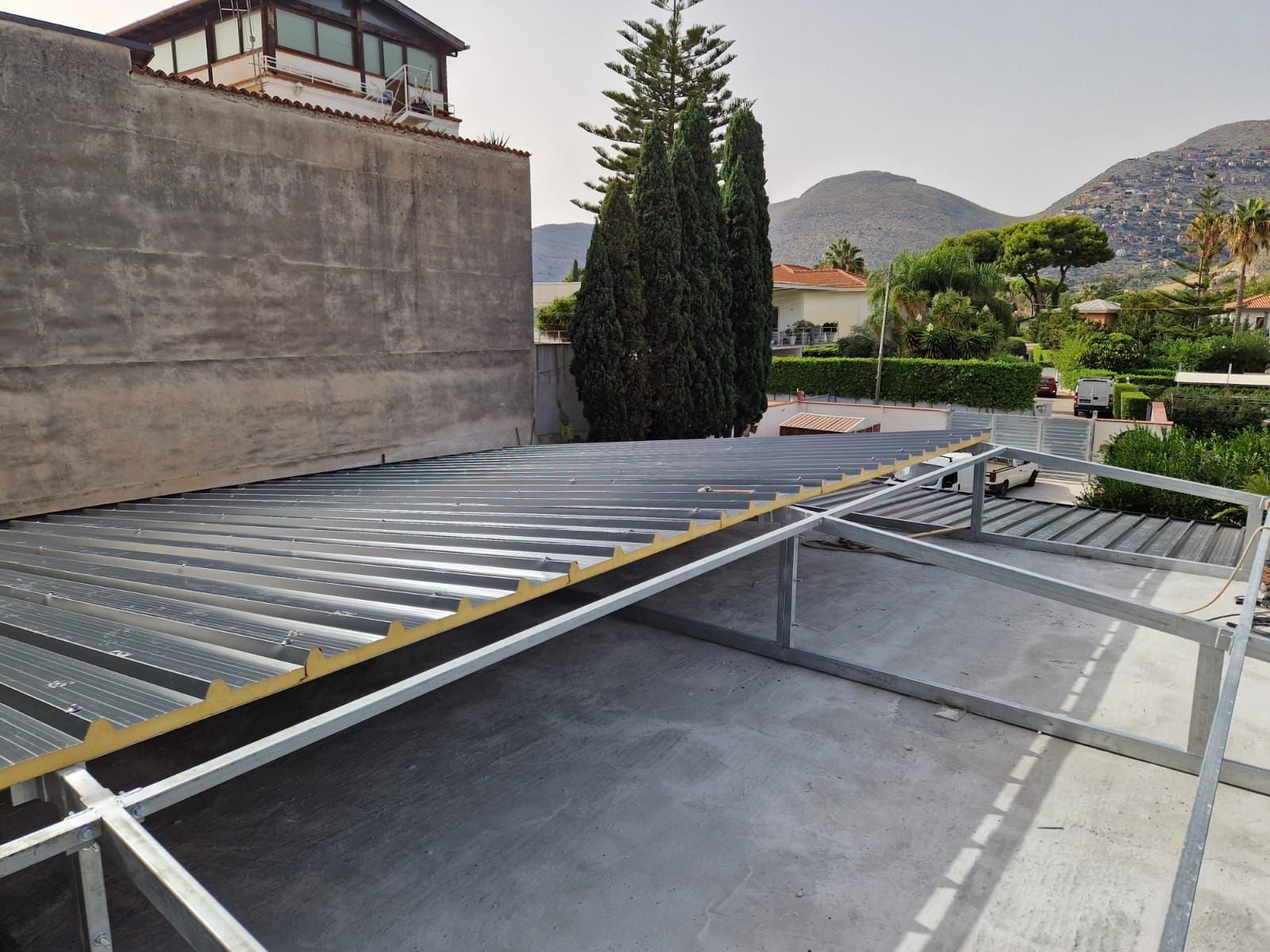 Impianto di pannelli solari installato su un tetto piano, accanto a un alto muro e ad alberi, con le montagne sullo sfondo.