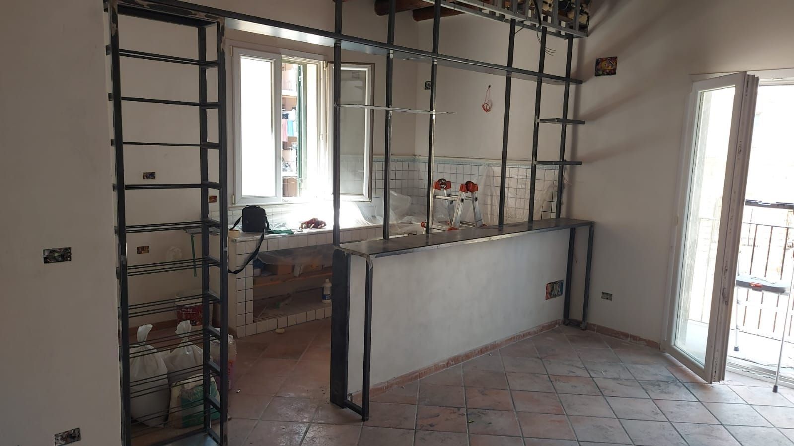 Camera parzialmente ristrutturata con struttura in metallo, pavimento piastrellato, finestra e porta finestra che si apre sul balcone.