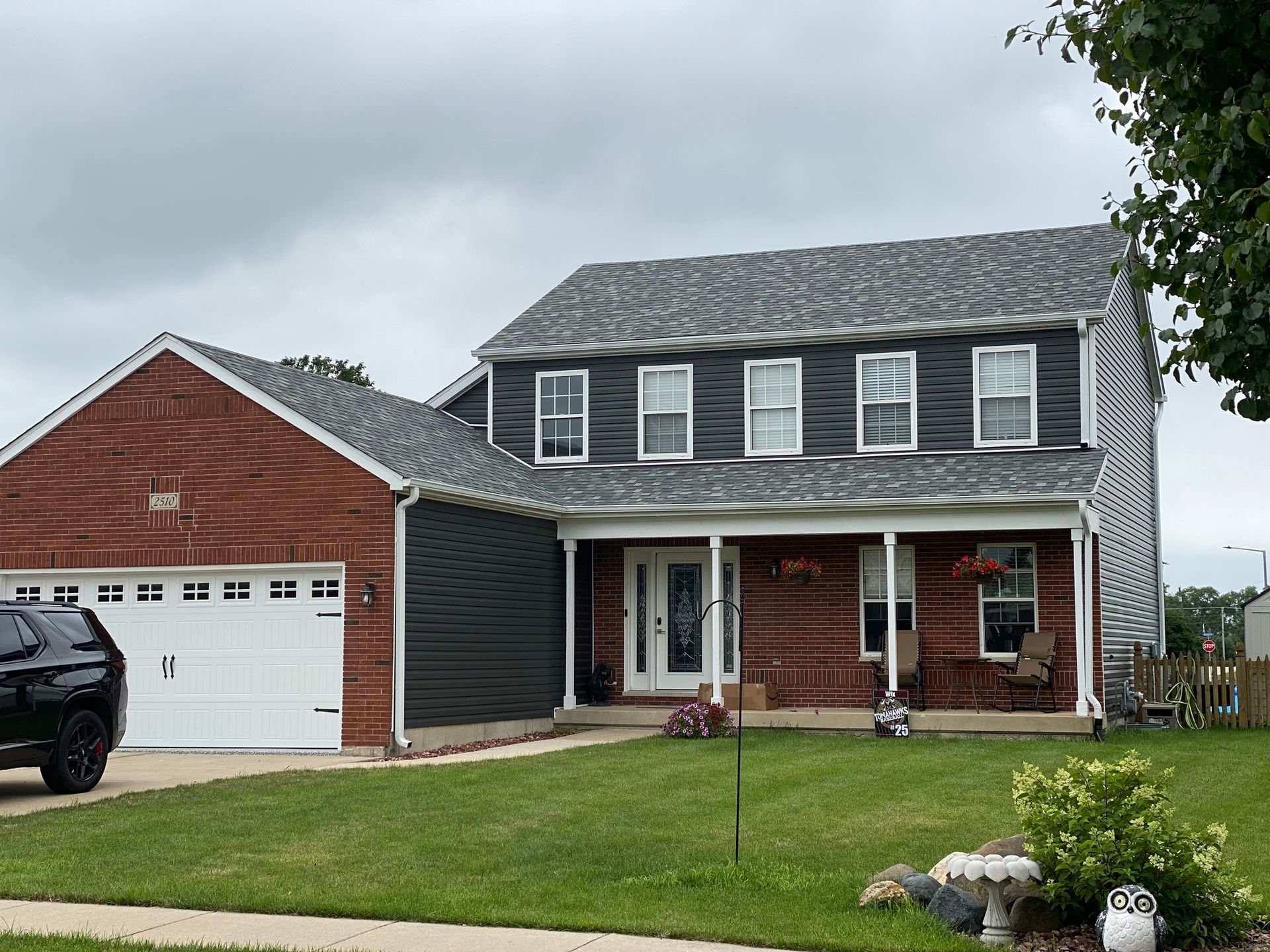 Suburban two-story house with brick and siding, front porch, garage, and green lawn under cloudy sky