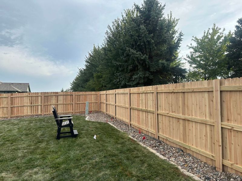 Backyard with wooden fence, grass lawn, black chair, and large evergreen trees under a cloudy sky