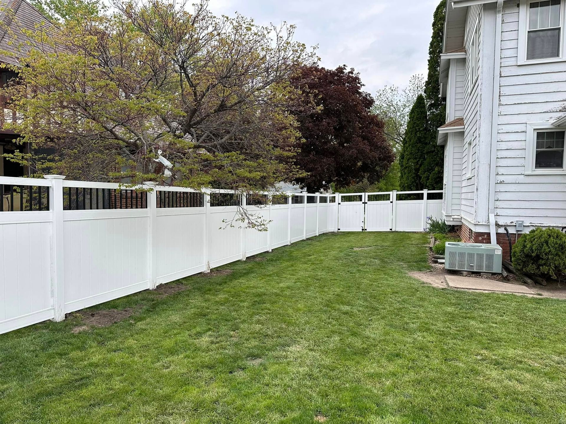 Backyard with green lawn, white fence, and a white house beside trees in bloom