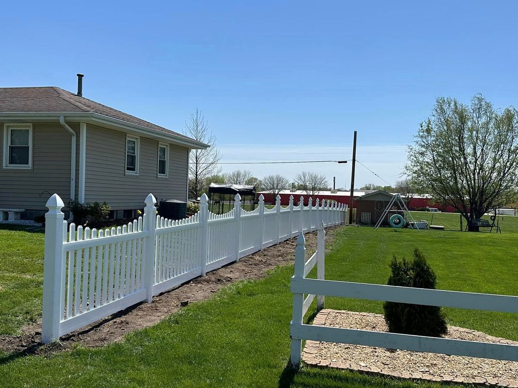 White picket fence beside a beige house and green lawn under a clear blue sky