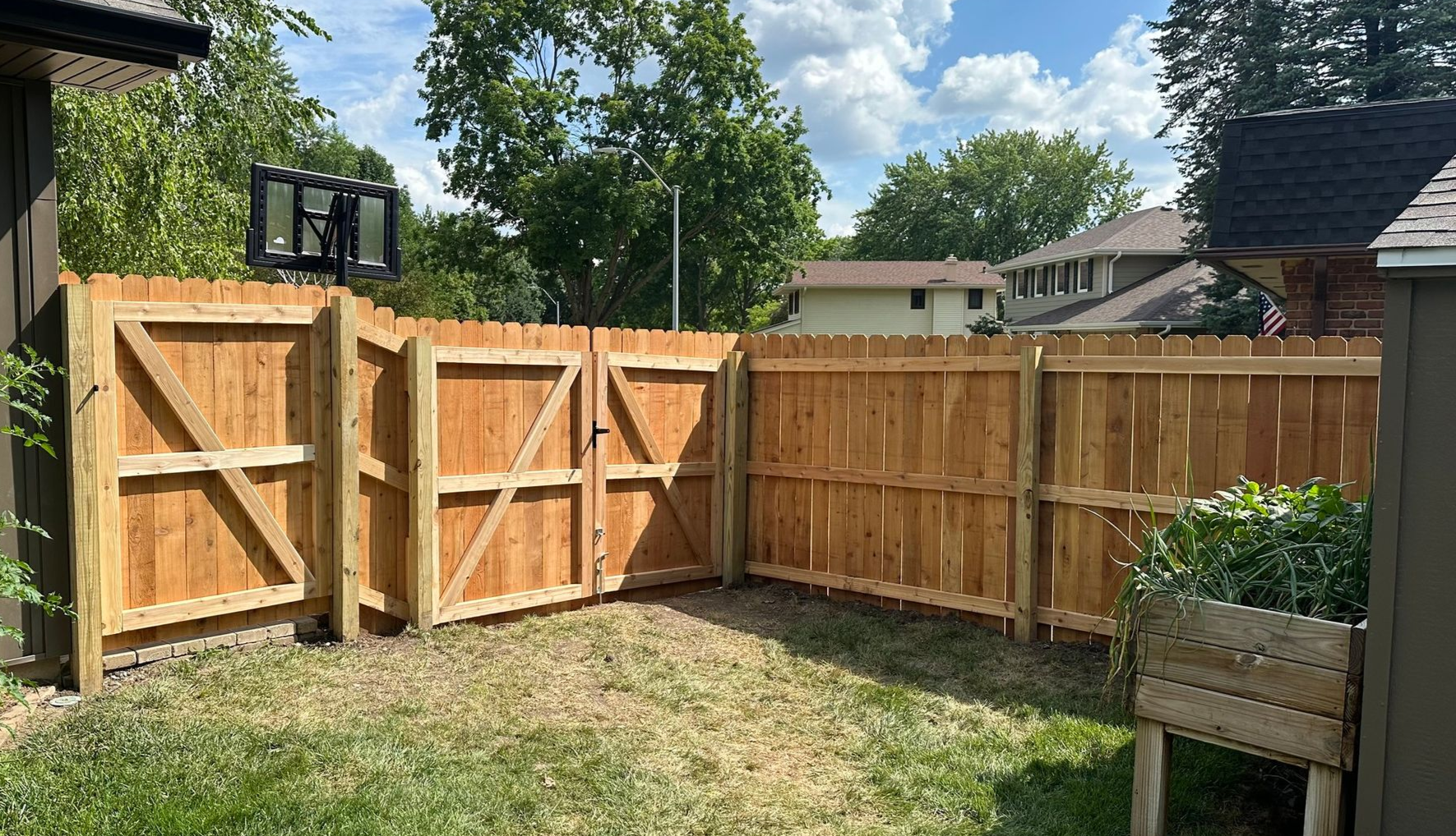 Wooden backyard fence with an open gate, grass lawn, and flower planter under a sunny sky