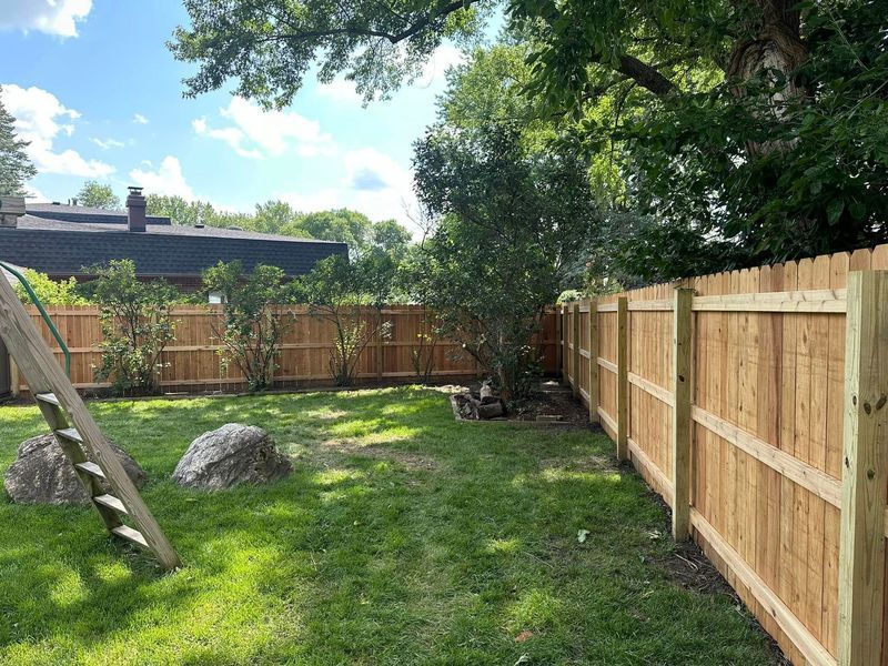 Backyard with wooden fence, ladder, and large rock under leafy trees on a sunny day