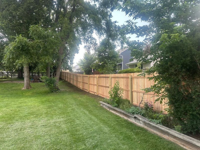 Tree-lined backyard with green lawn, wooden fence, and a raised garden bed along the right side.