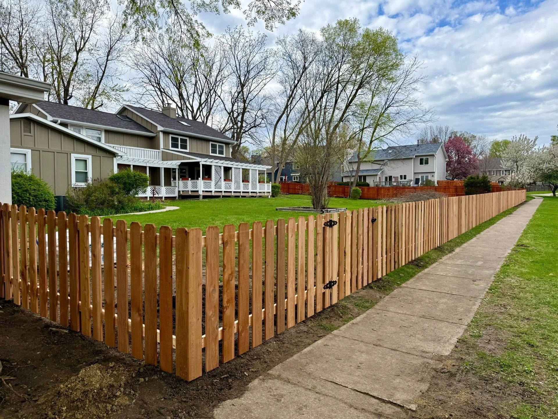 Wooden picket fence around a green yard beside a sidewalk and suburban houses