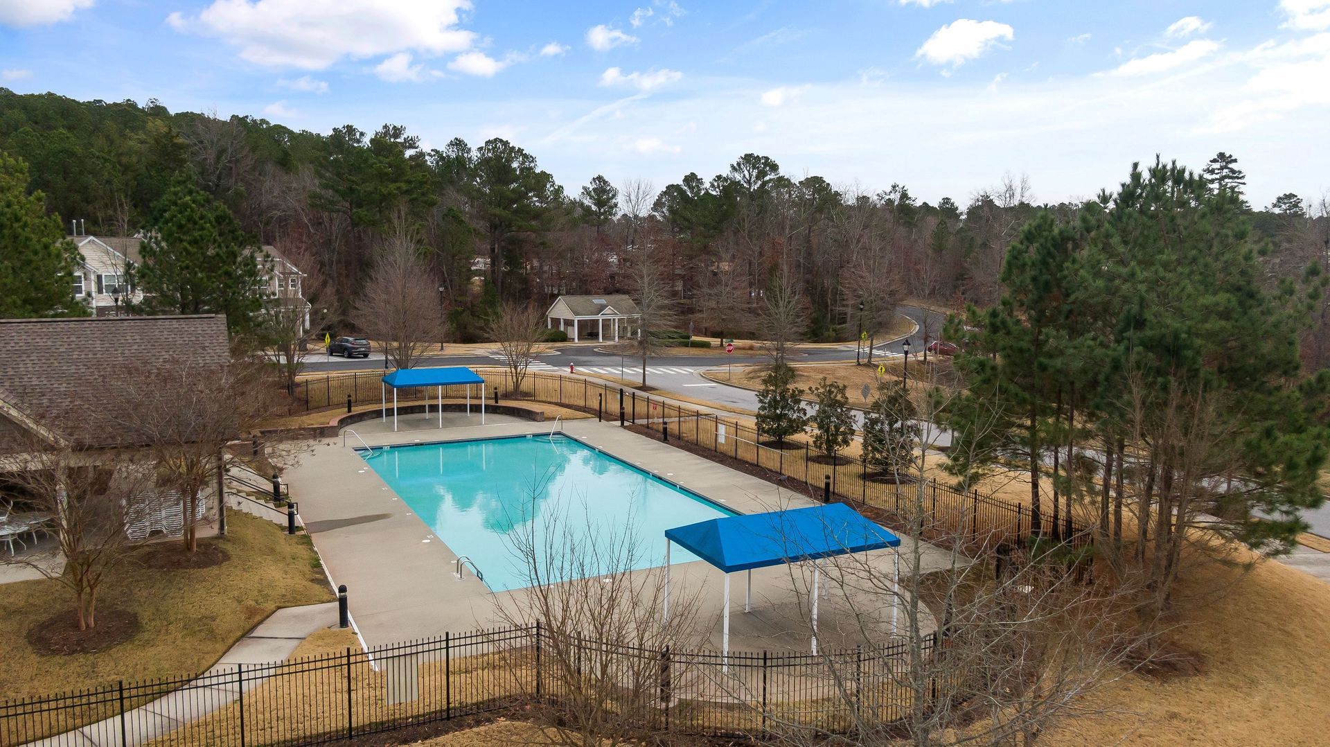 Outdoor swimming pool with blue shade covers, surrounded by a fence and lounge area under a cloudy sky