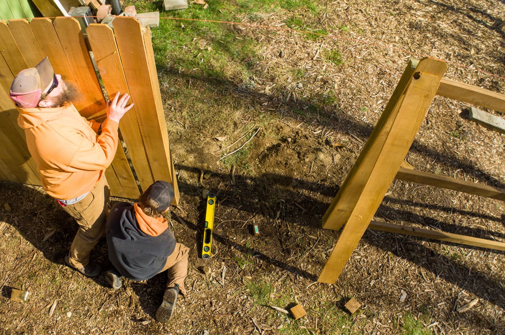 Two workers installing a wooden fence in a sunny yard, viewed from above.