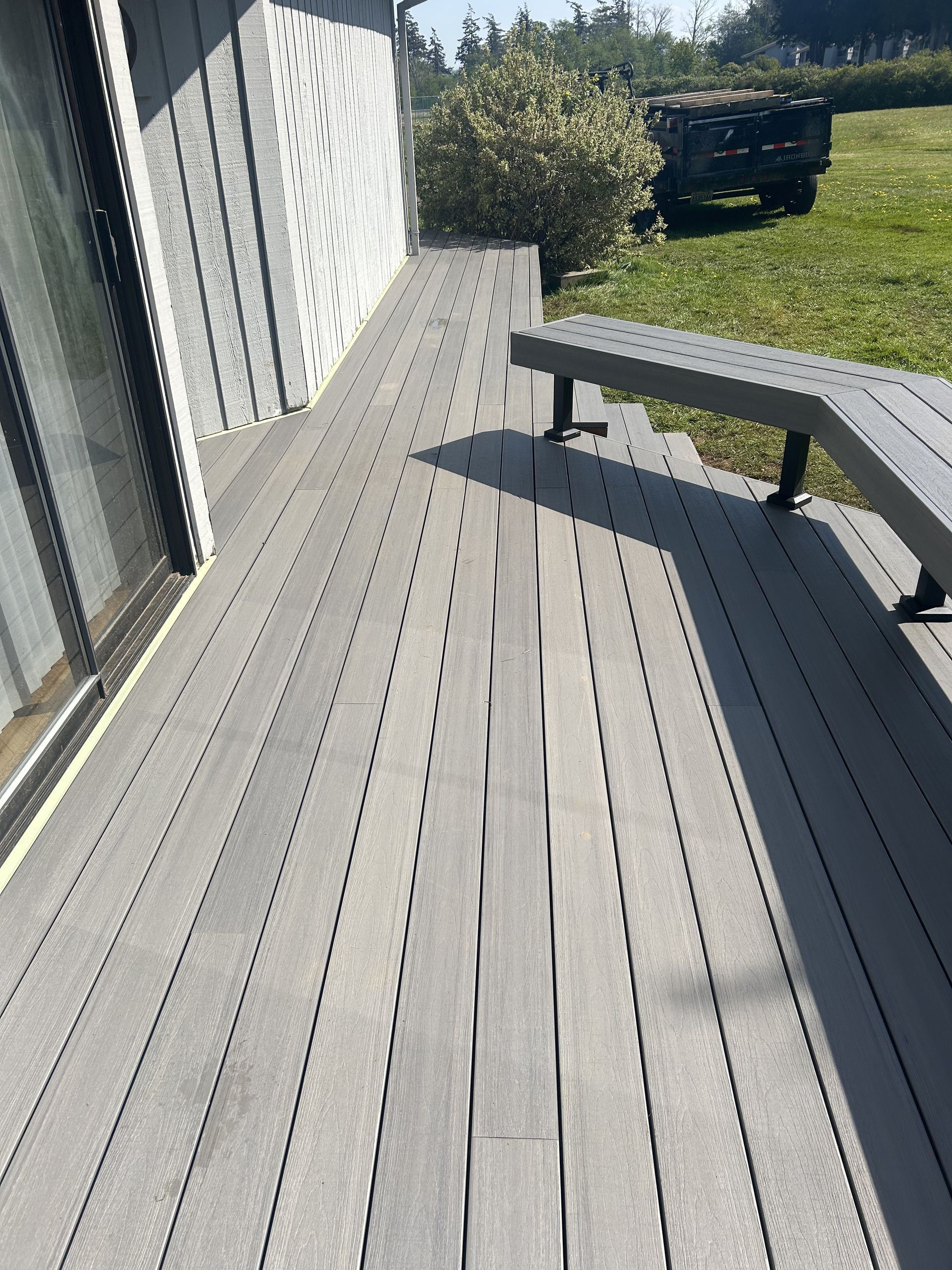 Gray wooden deck beside a house, with a low bench and grassy yard in sunlight.
