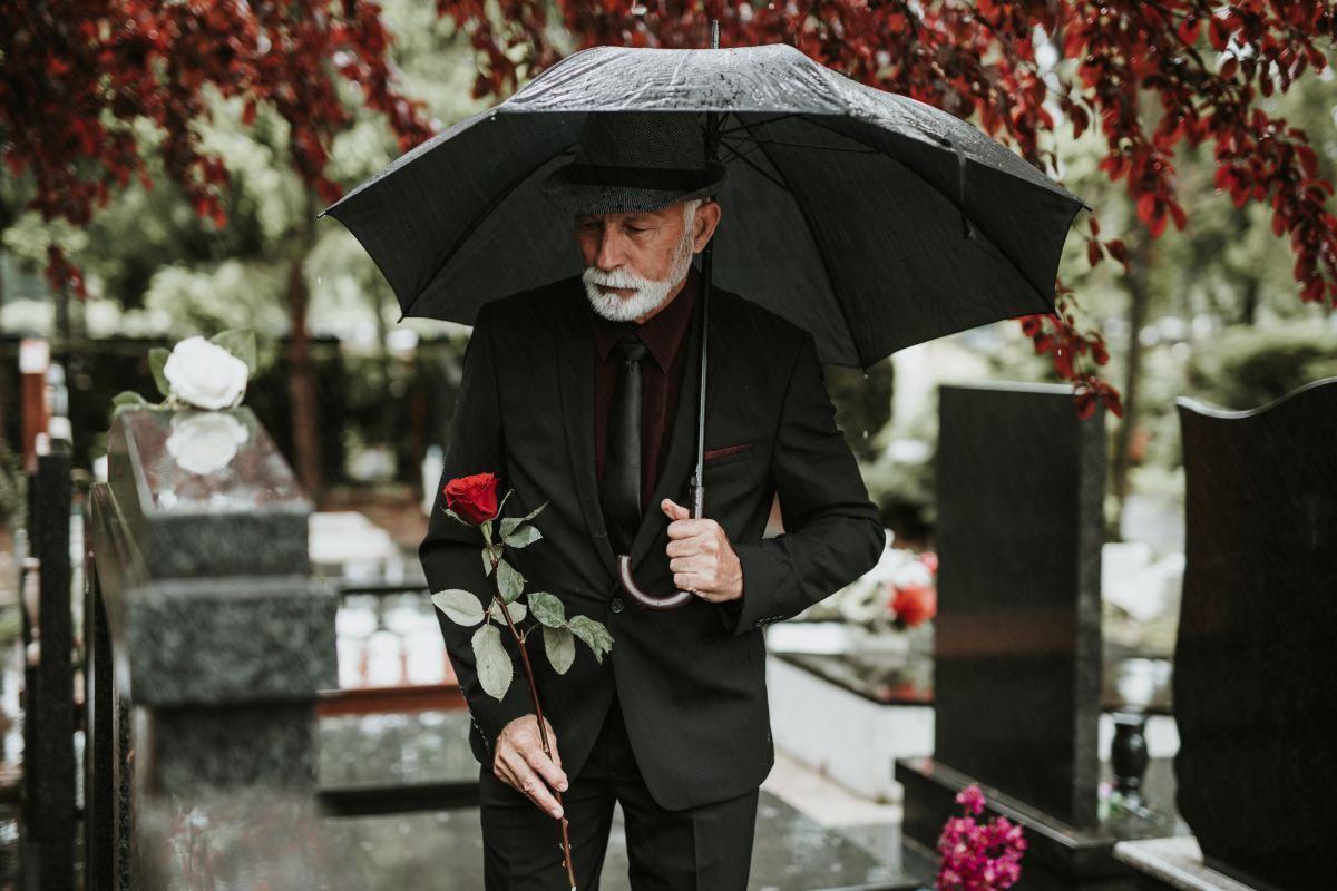 Grieving man in black suit at cemetery, holding umbrella and red rose, near tombstones.