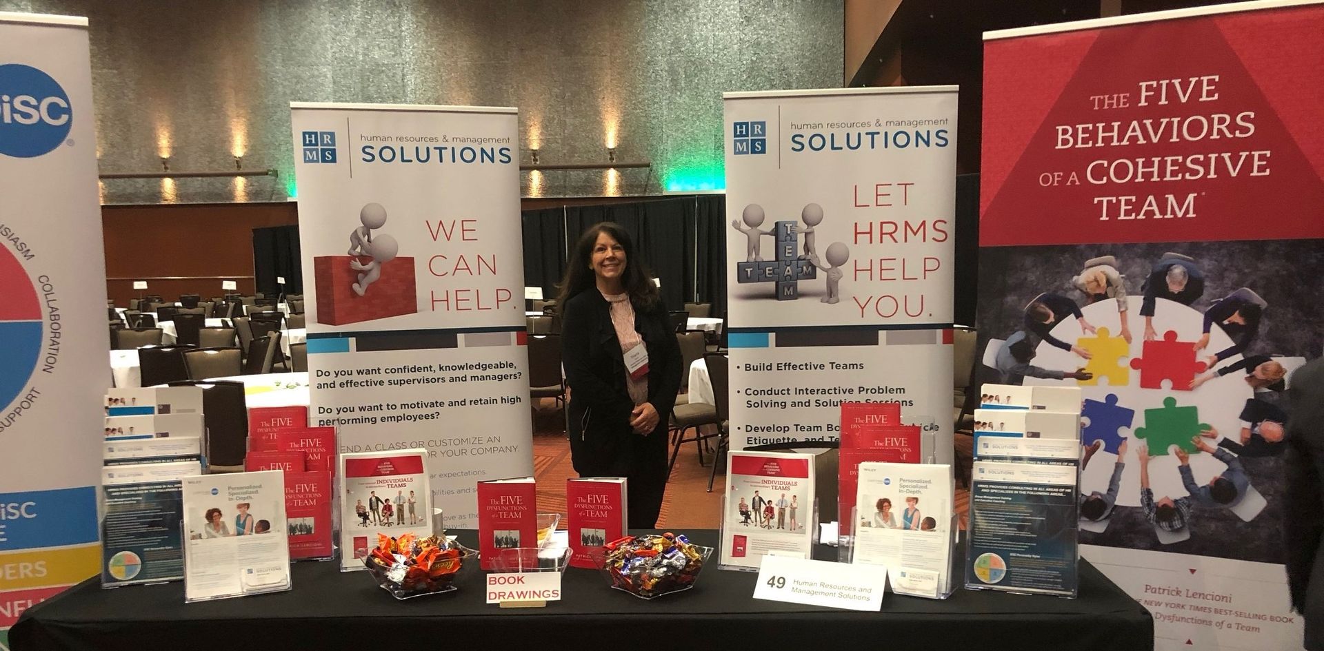 A woman stands in front of a table with a sign that says a five behaviors cohesive team