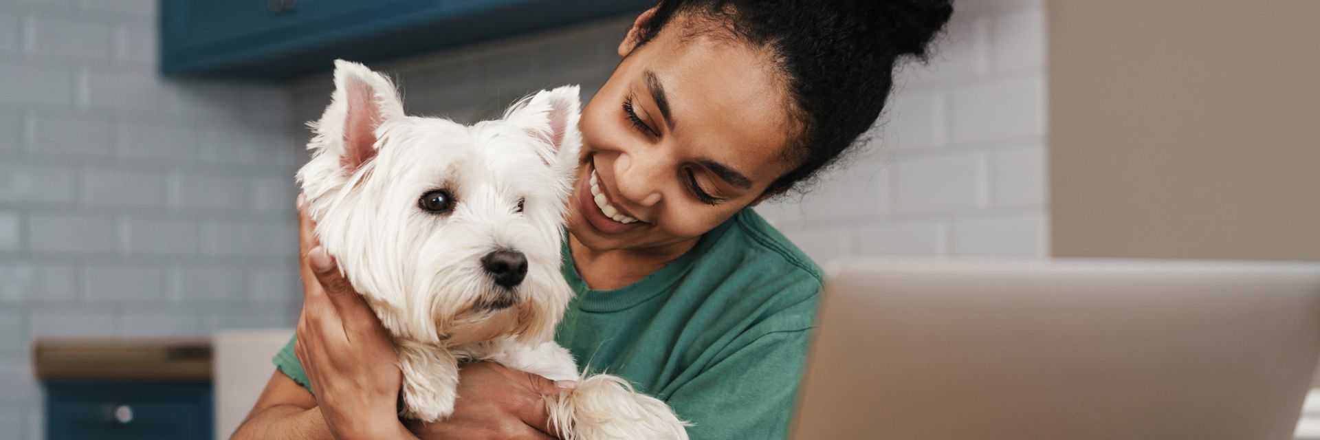 Woman smiling while cuddling a small white dog beside a laptop