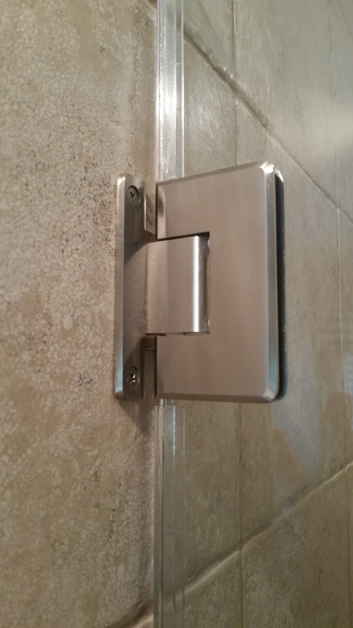 A close up of a stainless steel shower door hinge on a tiled wall