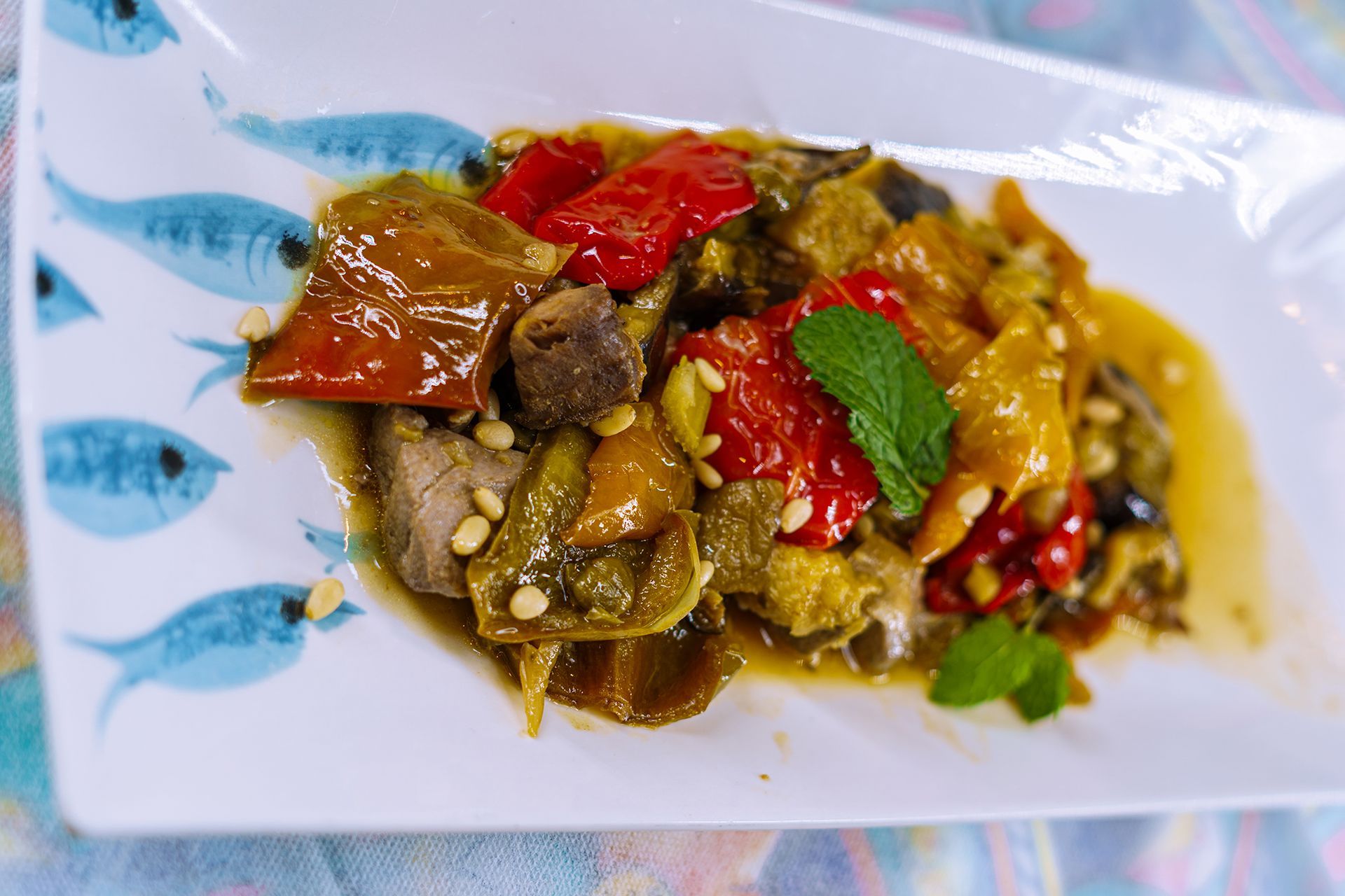 Misto di verdure con peperoni rossi, arancioni e verdi, su un piatto bianco decorato con pesci azzurri, guarnito con menta e pinoli.