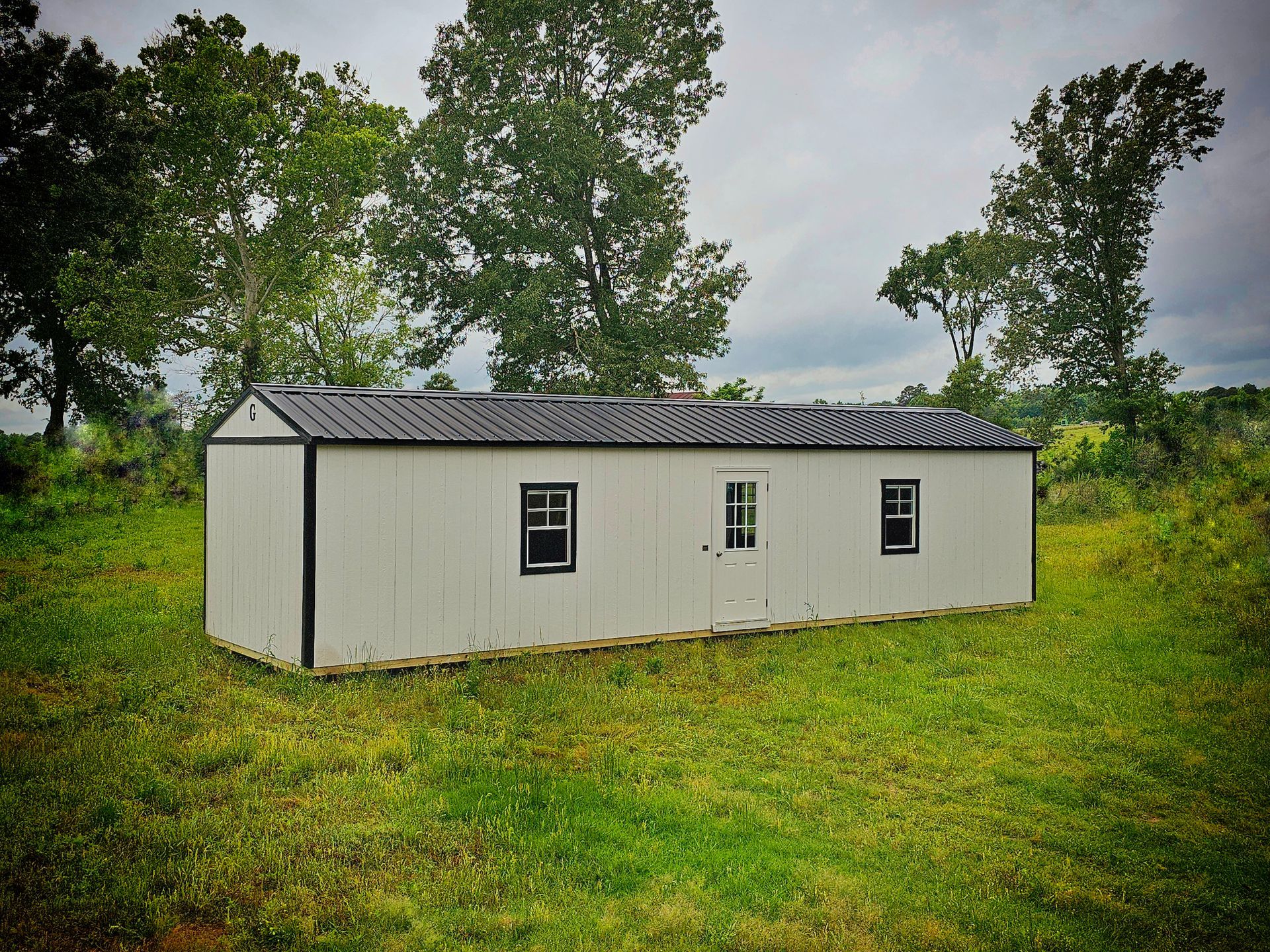 A white shed with a black roof is sitting in the middle of a grassy field.