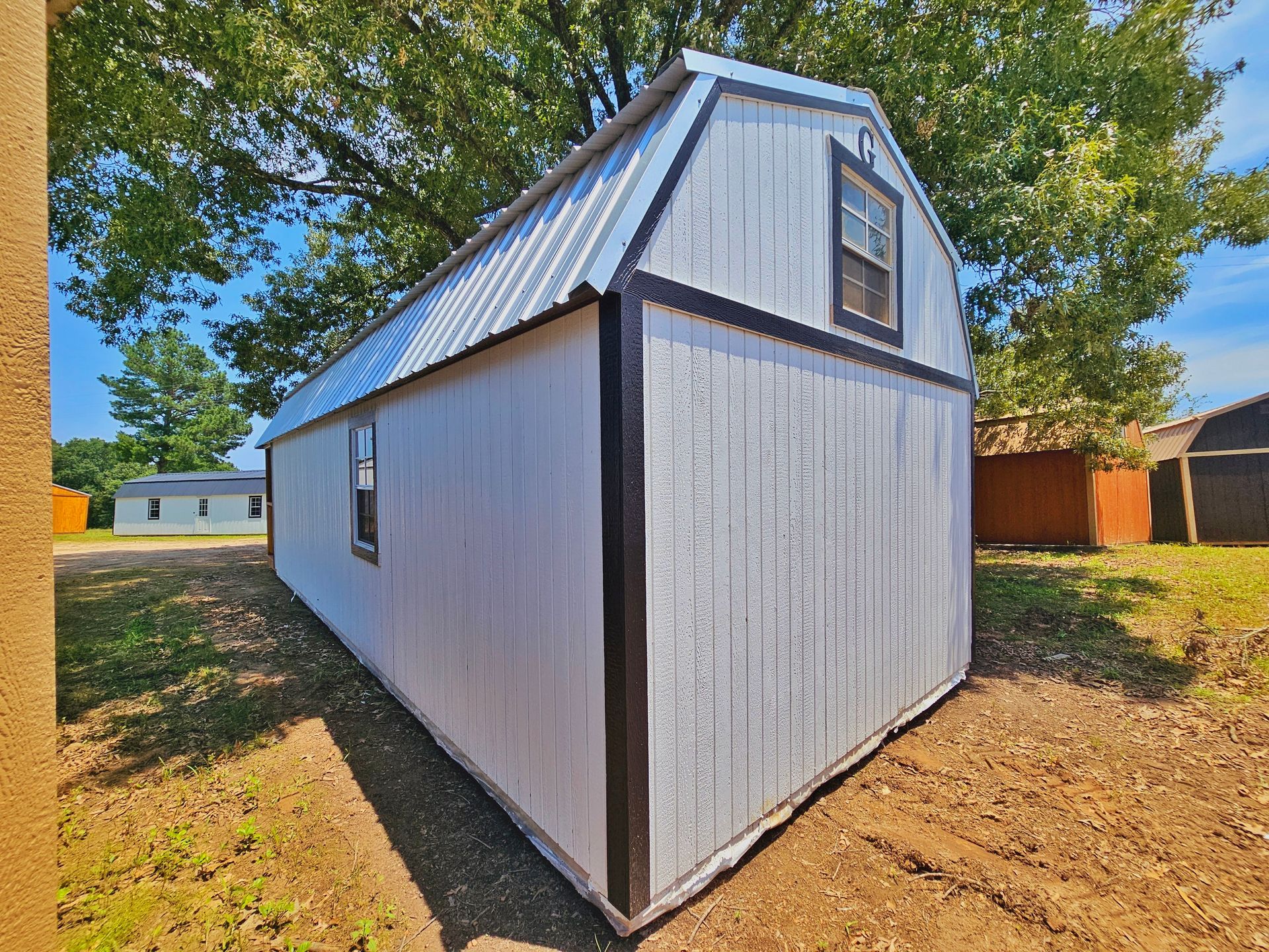 A white barn with a black trim is sitting in a dirt field.