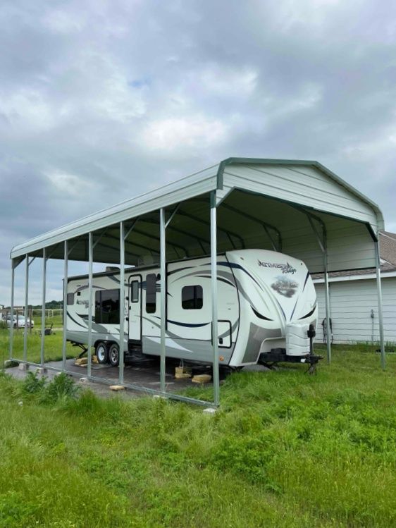 A white rv is parked under a carport in a grassy field.