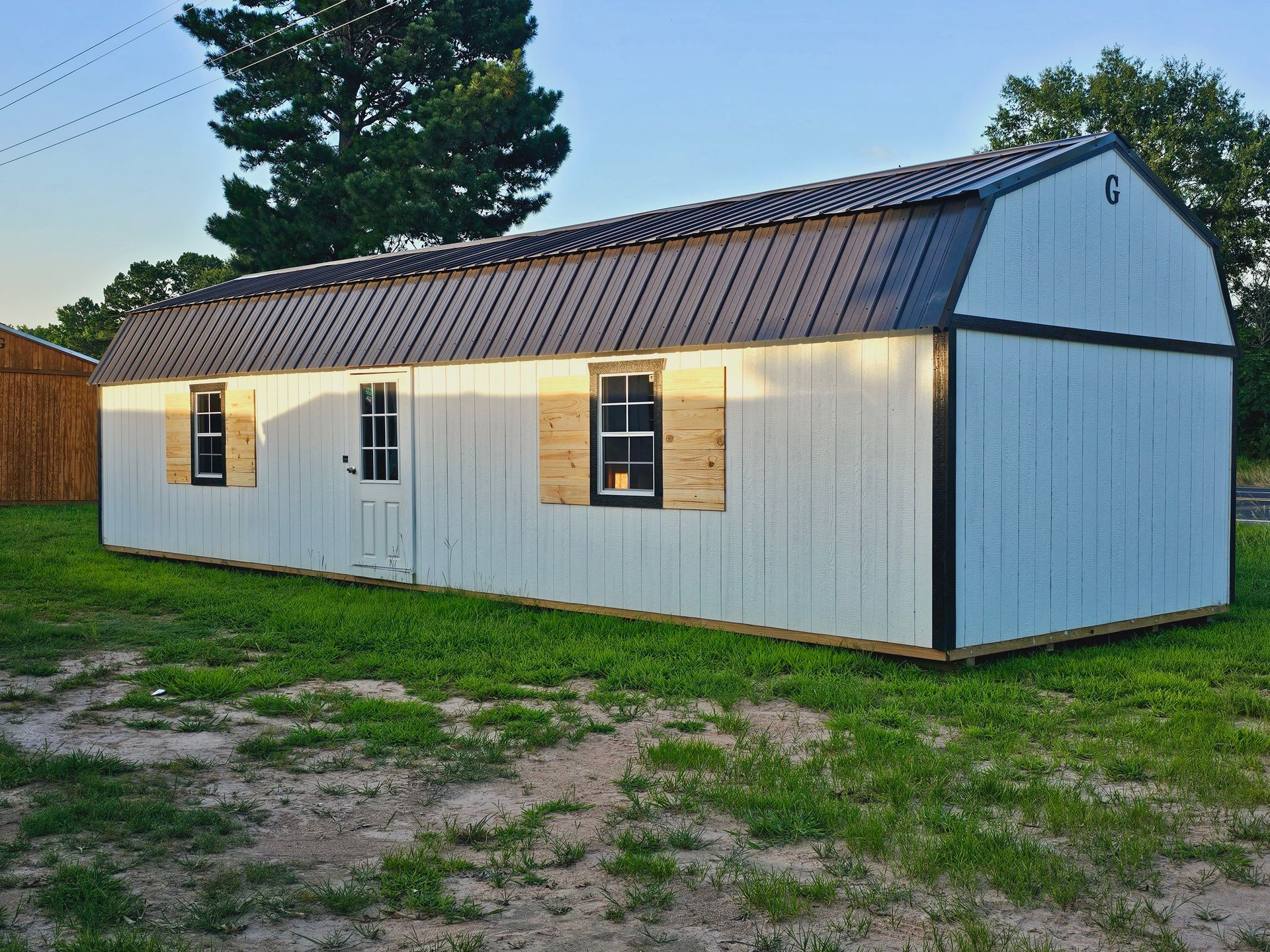 A white barn with a brown roof is sitting in the middle of a grassy field.