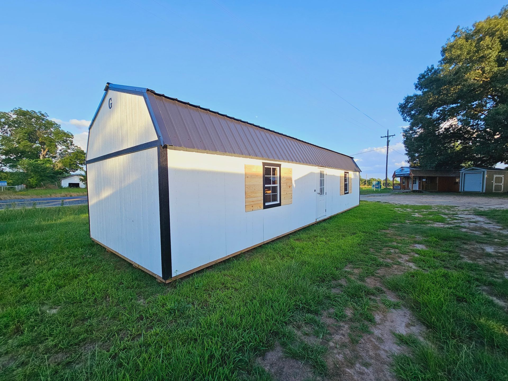 A white barn with a brown roof is sitting in the middle of a grassy field.