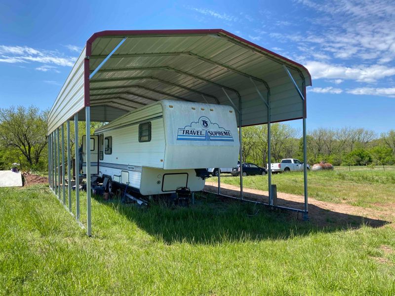 A white rv is parked under a metal carport in a grassy field.