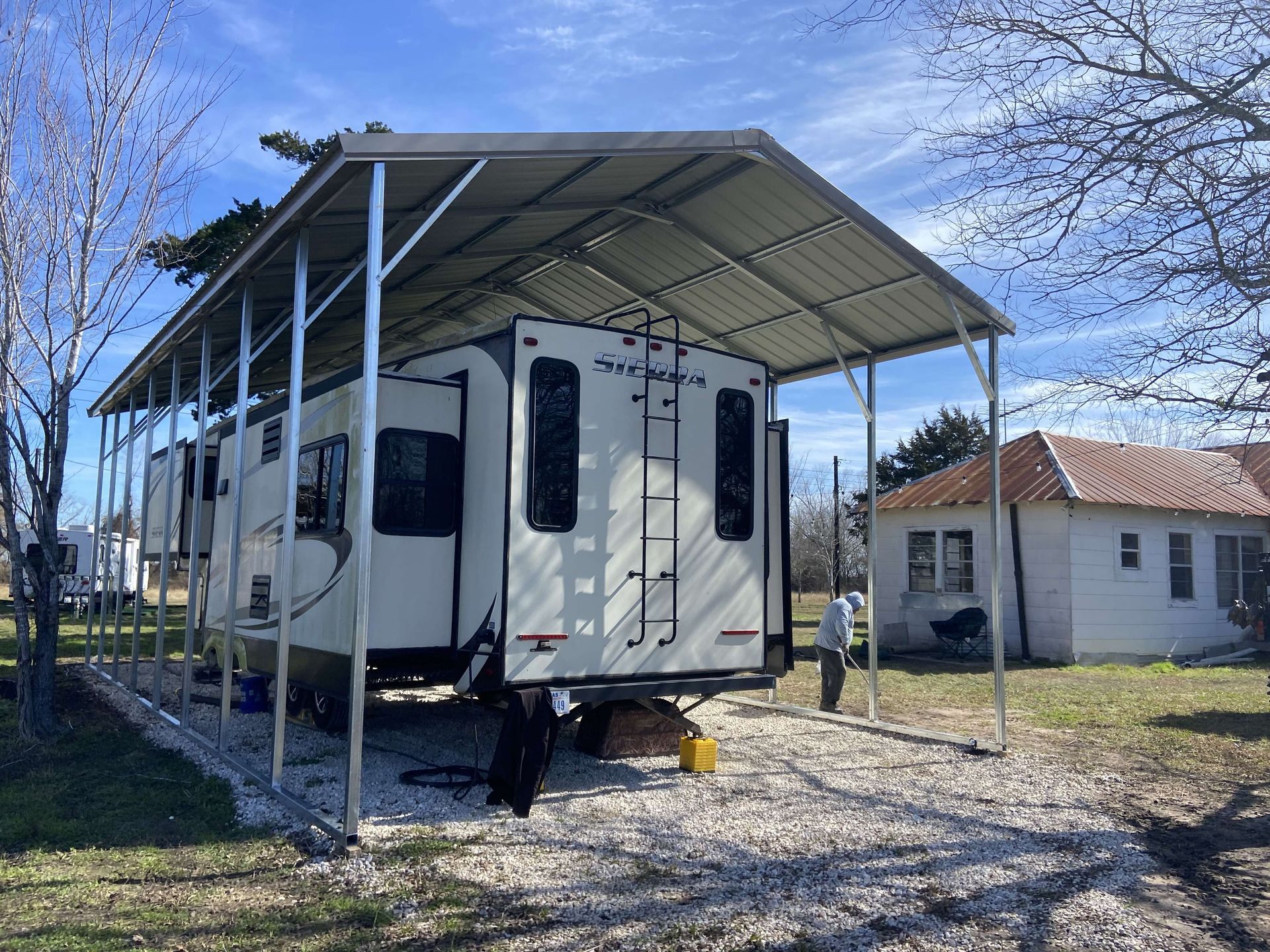 A white rv is parked under a metal roof in front of a house.
