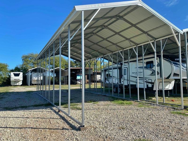 A rv is parked under a canopy in a gravel lot.