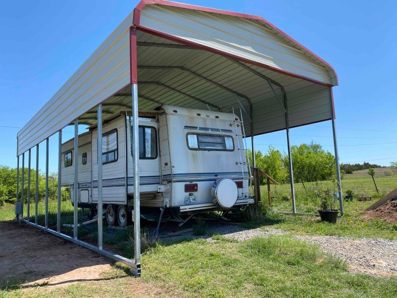 A rv is parked under a canopy in a field.