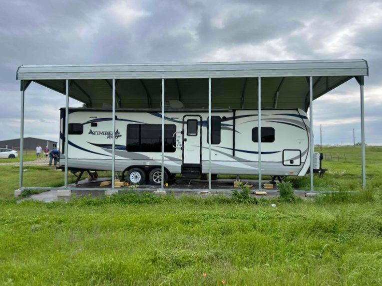 A rv is parked under a carport in a grassy field.