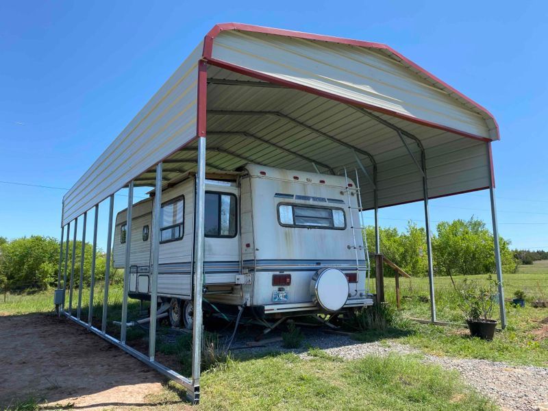 A white rv is parked under a carport in a field.