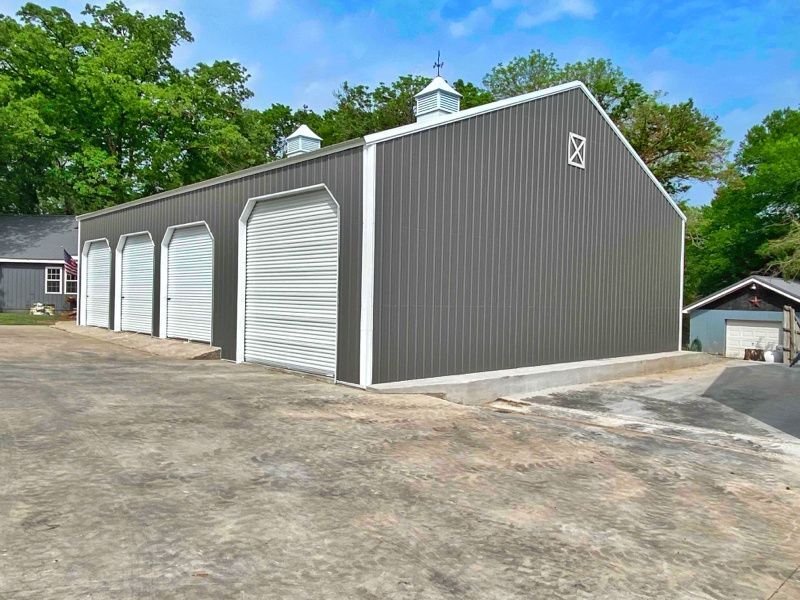 A large gray garage with white doors is sitting next to a house.