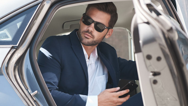 Man in suit exits car, holding phone, wearing sunglasses.