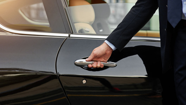 Person in a suit opening a black car door with a chrome handle; light interior visible.