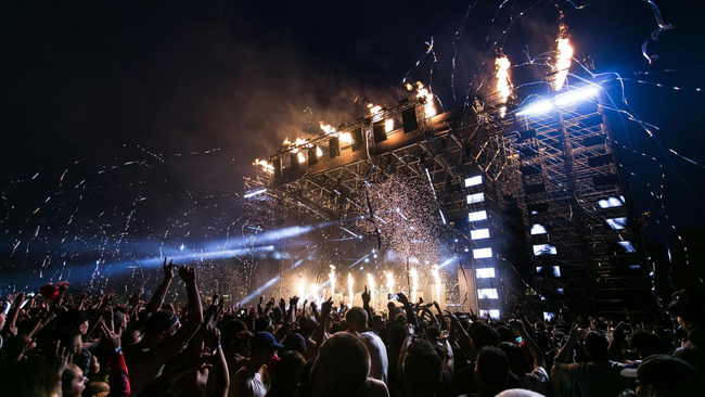 Concert crowd with stage, pyrotechnics, and confetti at night.