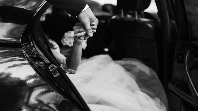 Bride in white dress and heart-shaped sunglasses smiles from vintage white car, holding flowers.