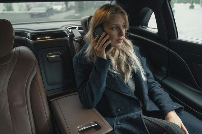 Woman in a car, talking on her phone, wearing a dark coat, looking concerned.