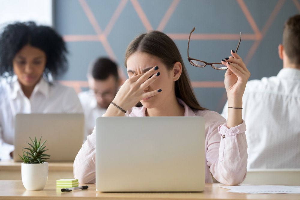 A Woman Is Rubbing Her Eyes While Sitting In Front Of A Laptop Computer — Wegner Therapies In Pialba, QLD