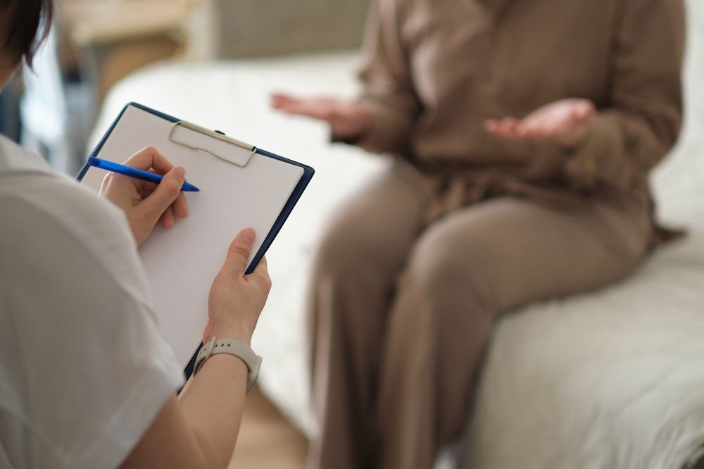 A Woman Is Sitting On A Couch Talking To A Doctor While Holding A Clipboard — Wegner Therapies In Pialba, QLD