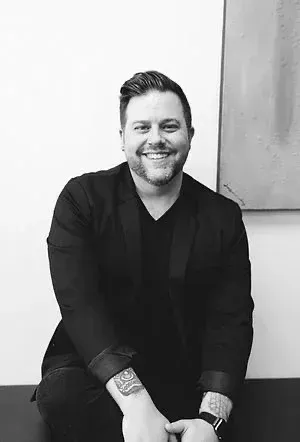 Man with tattoos smiling, wearing black blazer and shirt. Leaning against a wall.
