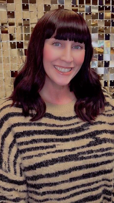 Woman with dark hair and bangs, smiling, in a zebra-striped sweater, against a mirrored wall.