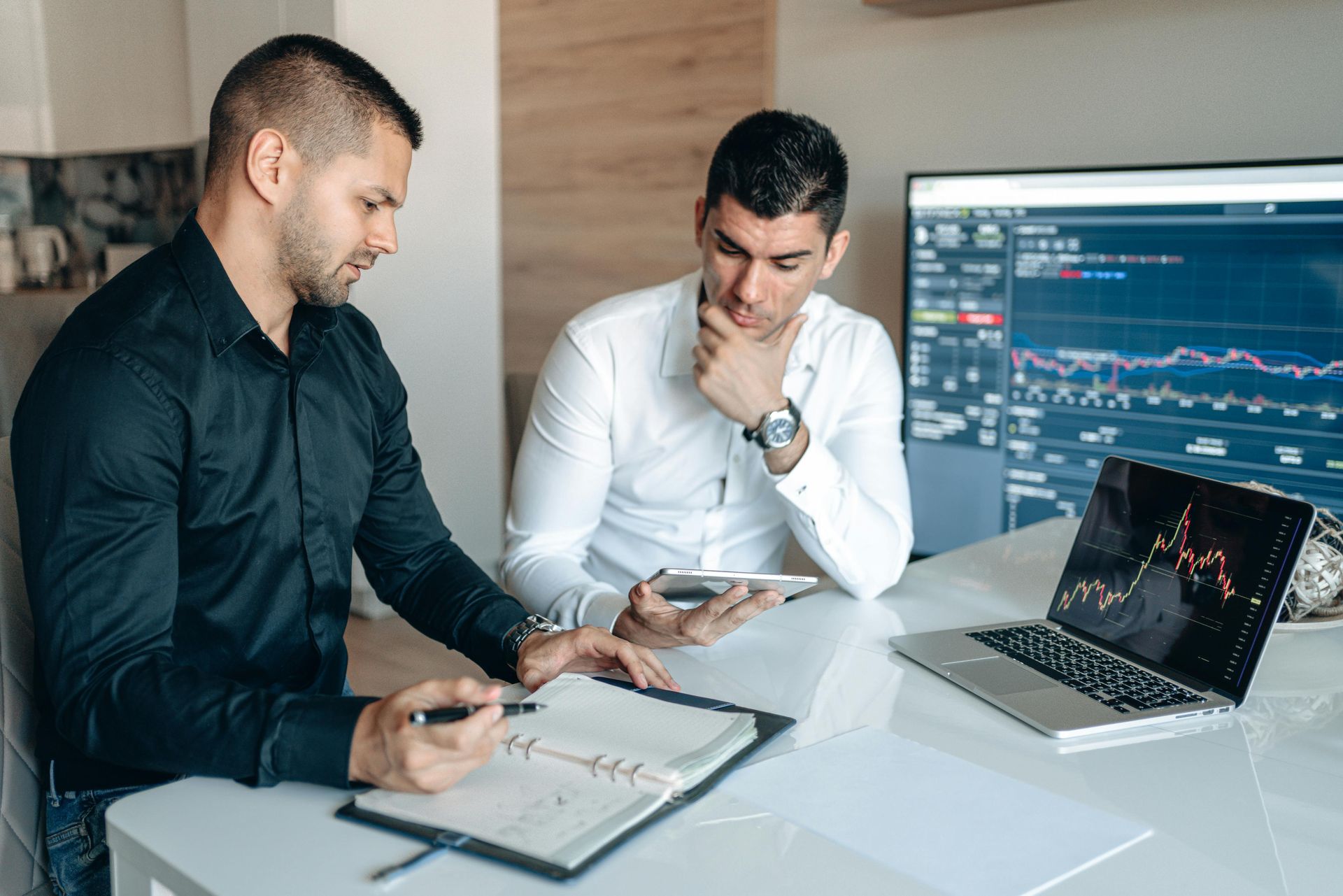 Two men at a table analyzing data on a laptop, tablet, and large screen. They look focused, possibly in an office setting.