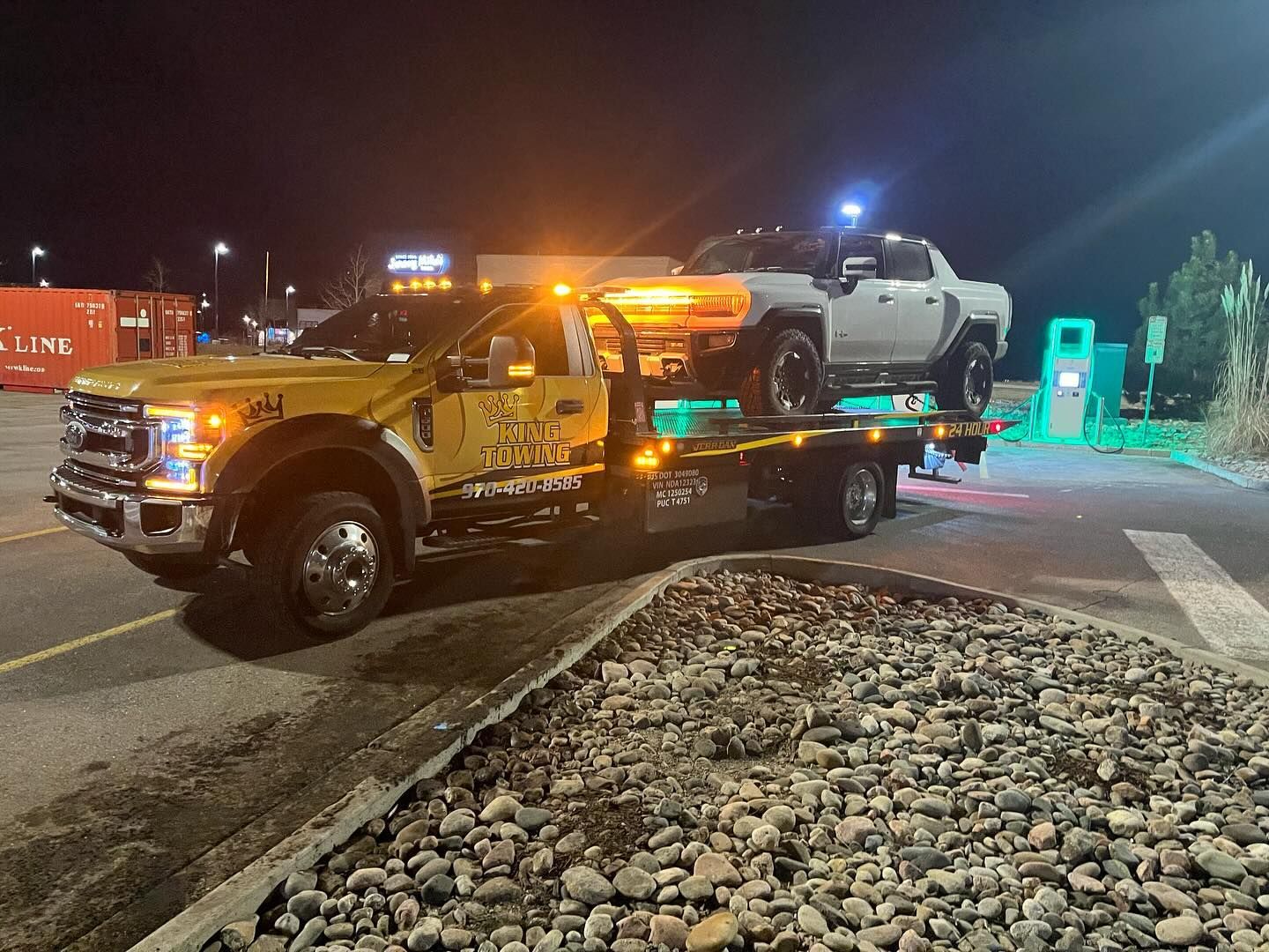 Tow truck hauling a white pickup at night in a lit parking lot near a rocky median
