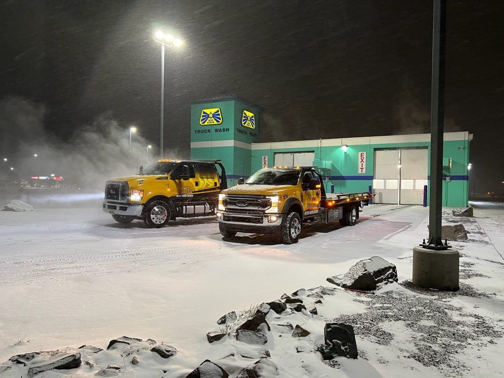 Two yellow tow trucks parked in a snowy lot beside a lit green building at night