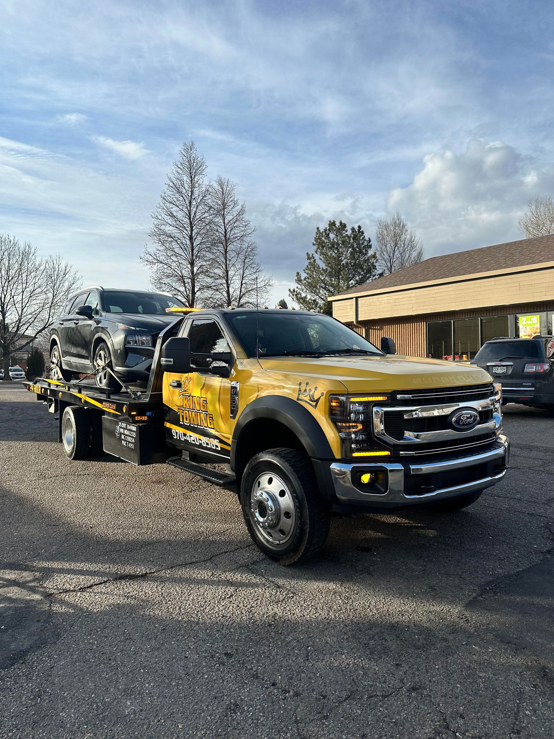 Yellow-and-black tow truck carrying a black car on a gravel lot beside a building