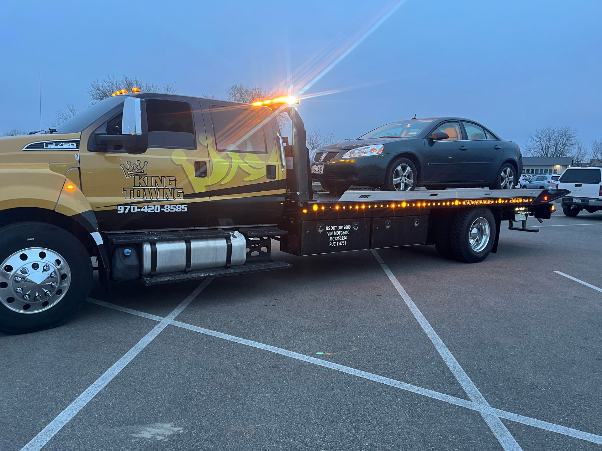 Tow truck hauling a gray sedan in a parking lot at dusk, with flashing amber lights.