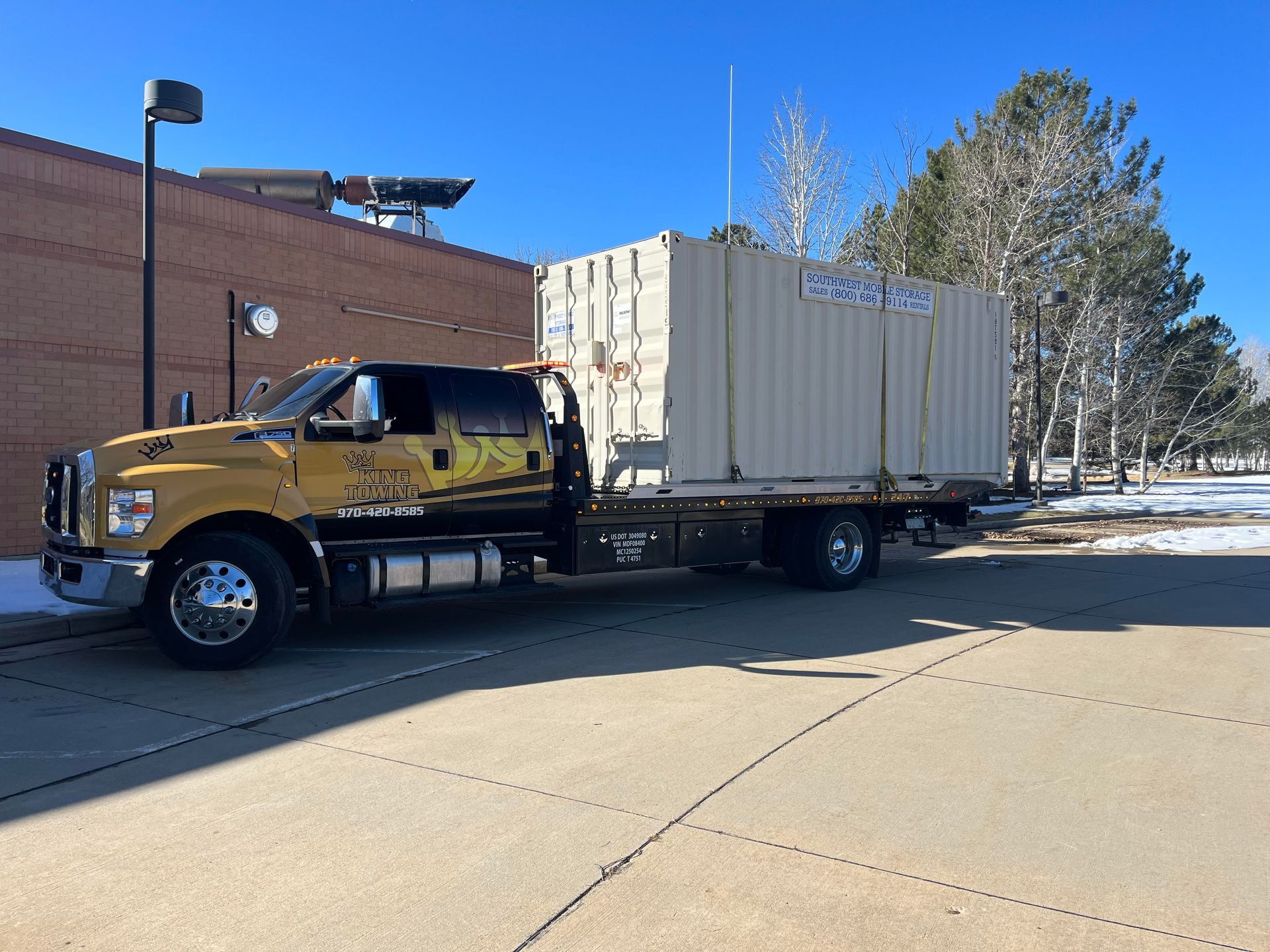 Yellow semi-truck with a white cargo container parked beside a brick building on a sunny street