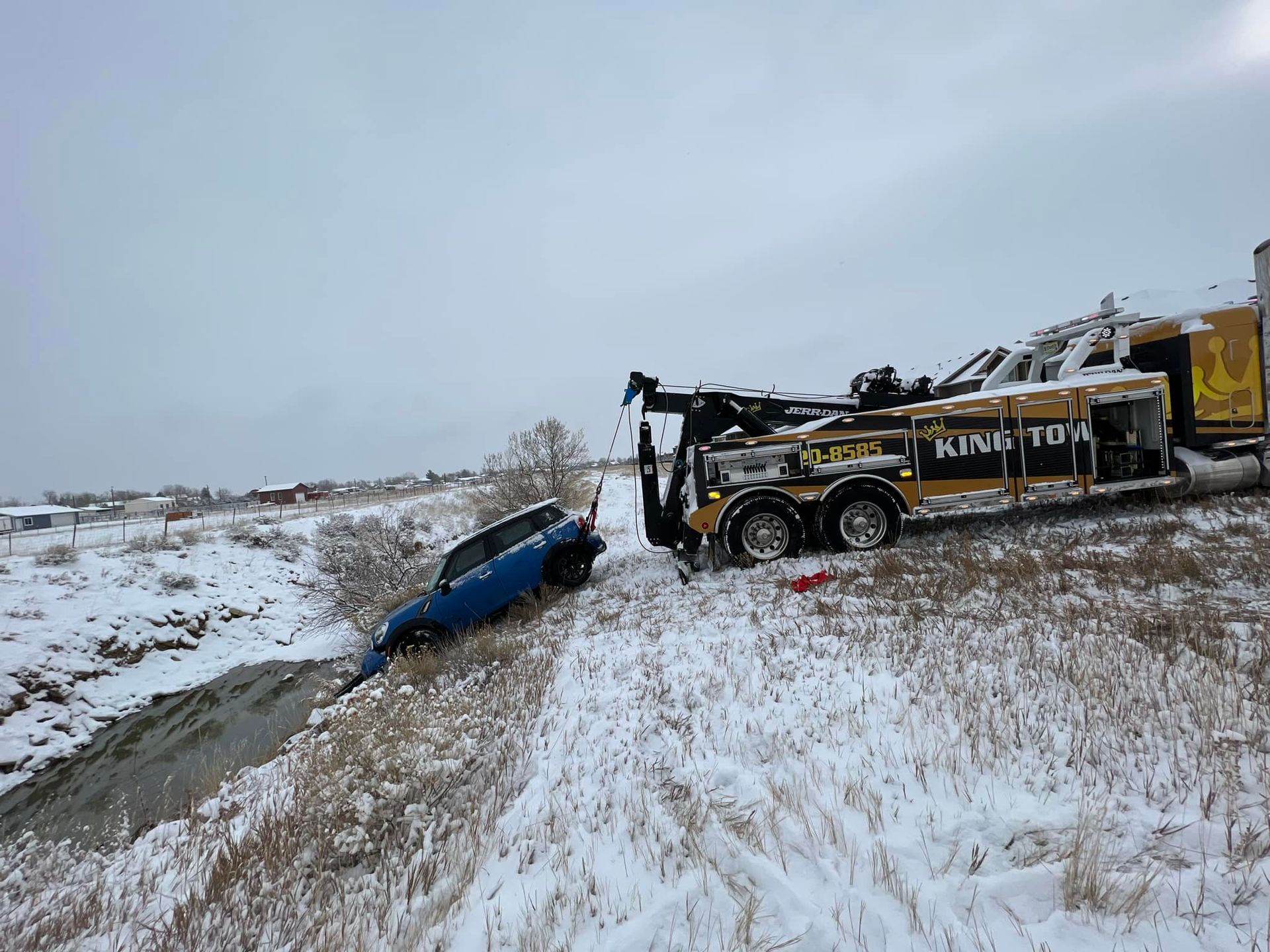 Tow truck pulling a blue car from a snowy ditch beside a road