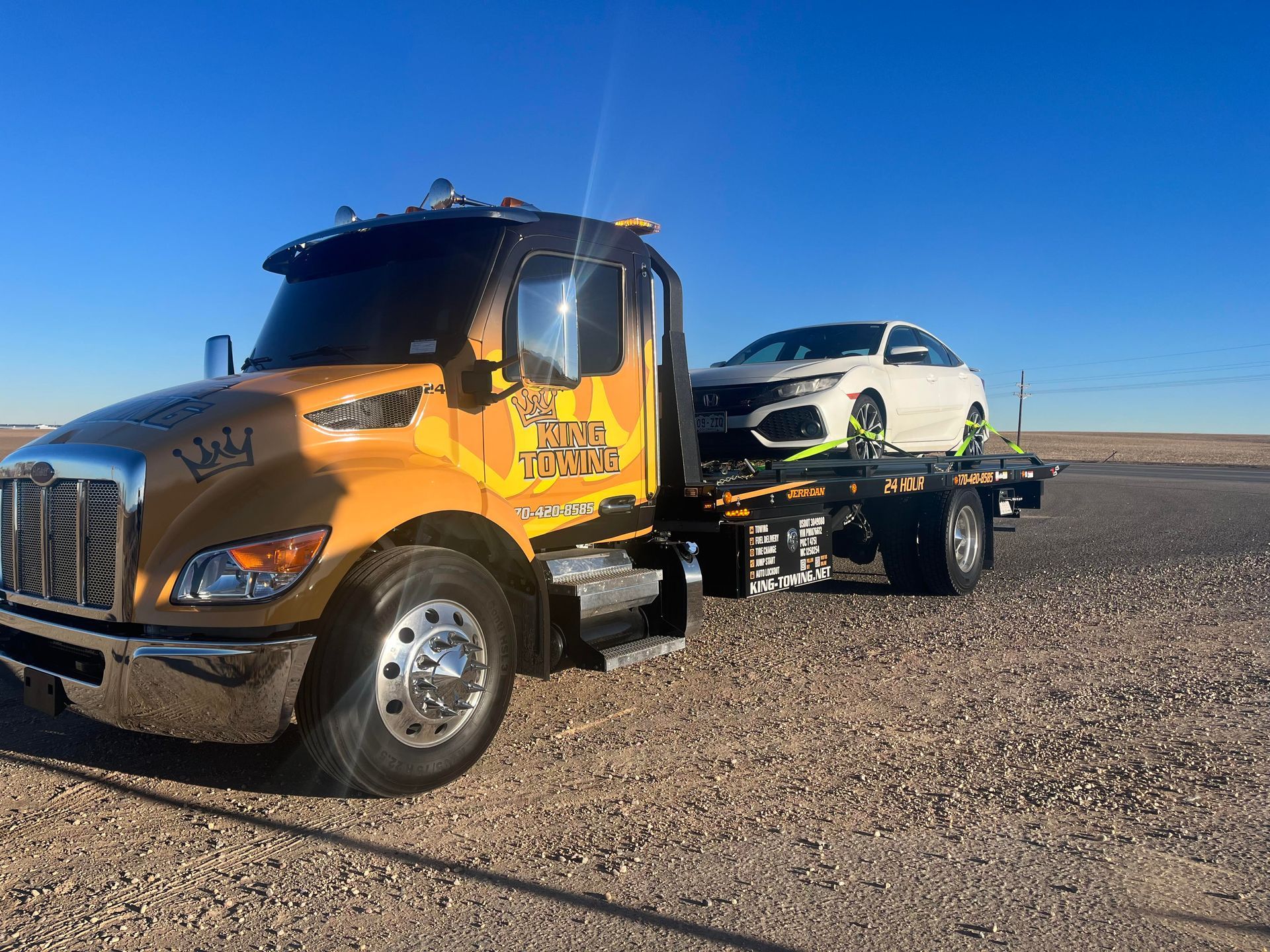 Yellow tow truck hauling a white car on a flatbed across a gravel lot under a clear blue sky