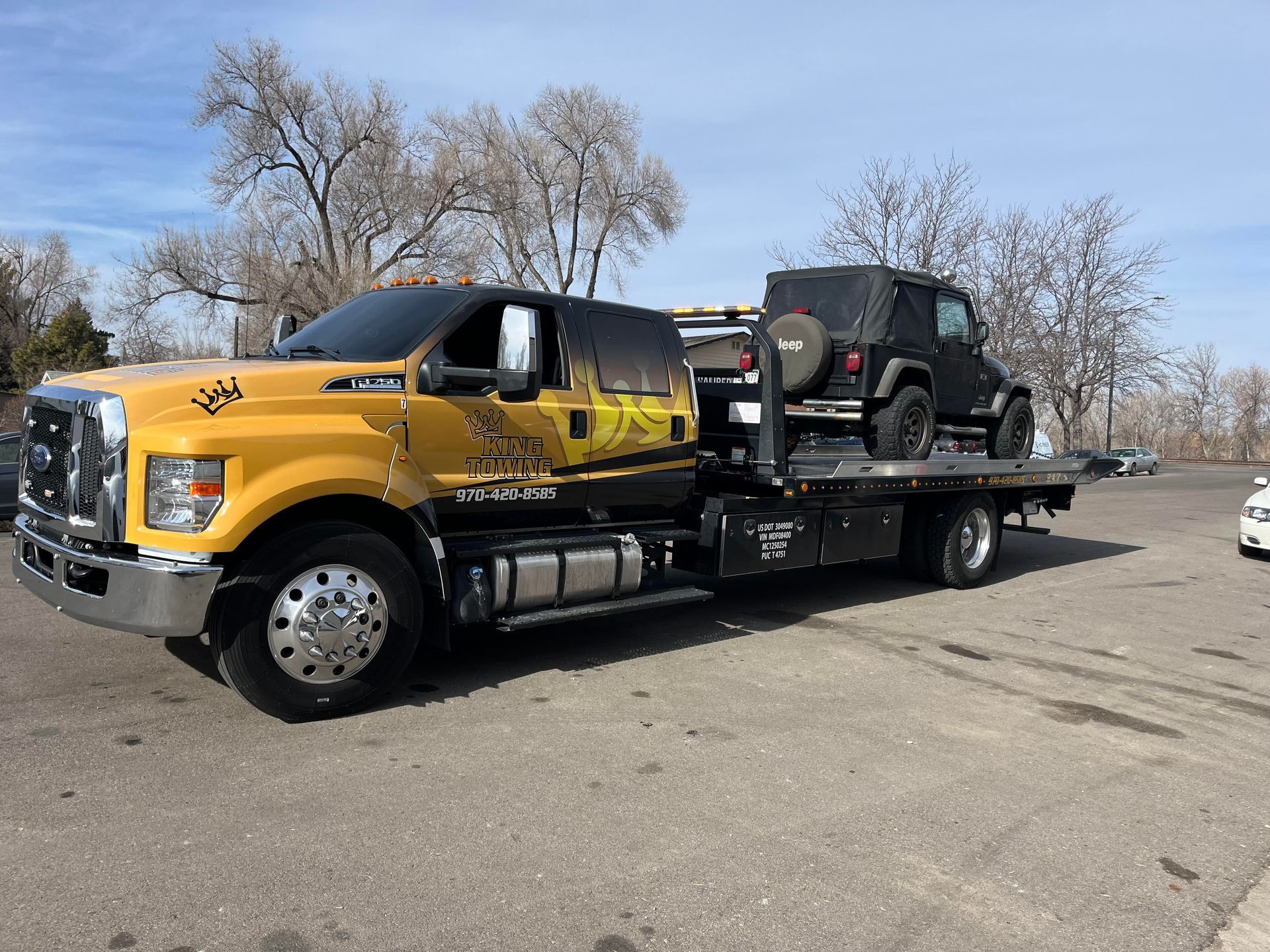 Yellow flatbed tow truck carrying a black Jeep on a roadside under a clear sky.